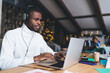 © BullRun - African American man working remotely on laptop, wearing headphones and focused. Cozy workspace with warm lighting, festive decorations. Background shows relaxed atmosphere and blurred holiday lights