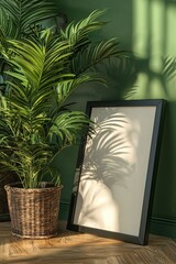 Poster - A minimalist interior scene featuring a blank frame beside a lush potted plant, with soft shadows cast on a green wall, perfect for home decor.