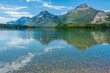 © CaptureNature - The Rocky Mountains reflect in the lake below it in Waterton Lakes National Park, Canada. The sky is a gorgeous blue, with white clouds. The Prince of Wales Hotel is in the background, and colorful ro