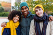 © Xavier Lorenzo - Three young diverse friends in warm clothes smiling at camera standing together outdoors. Gen Z student people standing at campus college. Education and friendship concept.