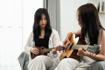 Two Asian women are playing guitars together in a living room. One of them is playing a ukulele