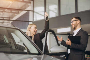 Wall Mural - Modern happy woman raising hand up making fist near her new car in car showroom. Bearded seller consultant in a suit