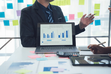 Business Analysis Meeting:  A professional businessman presenting financial data on his laptop, surrounded by sticky notes and other office supplies, to a colleague during a business meeting. 