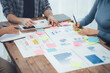 © Nittaya - Collaborative Brainstorming: A team of professionals gather around a table, their hands reaching for sticky notes, representing a dynamic exchange of ideas and strategies.