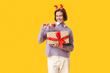 Wall Mural - Beautiful young happy woman in reindeer horns with gift boxes and shopping cart on yellow background. Christmas celebration