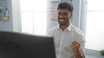 Young man smiling cheerfully in an office setting, with a clenched fist, sitting in front of a computer