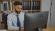 © Krakenimages.com - Handsome young hispanic man with a beard working at a computer in an office room with organized shelves in the background.