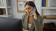 © Krakenimages.com - Young woman with brunette hair talking on a smartphone in an office workspace, appearing focused and engaged with bookshelves and a bulletin board in the background.