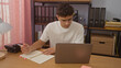 © Krakenimages.com - Young hispanic man taking notes while working on a laptop in an office with bookshelves in the background.