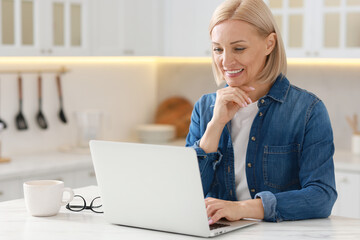 Wall Mural - Happy middle aged woman using laptop at white marble table in kitchen