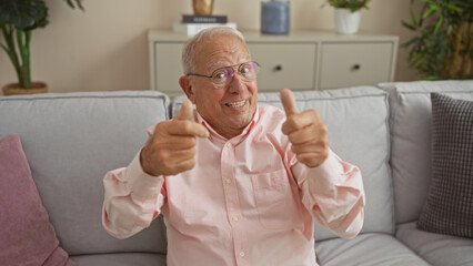 Happy, elderly, grey-haired, man in a pink shirt giving thumbs up while sitting on a couch in a cozy living room at home.