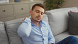 © Krakenimages.com - Handsome young hispanic man in a blue shirt with a beard sits in a modern living room, appearing tired and touching his neck, suggesting possible neck pain at home.