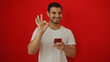 © Krakenimages.com - Hispanic man smiling while holding a smartphone and making an okay gesture against a vibrant red background, wearing a casual white t-shirt, isolated in a studio setting