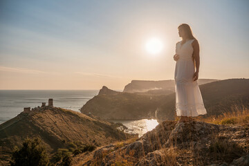 Wall Mural - A woman stands on a rocky hill overlooking the ocean. She is wearing a white dress and she is enjoying the view. The scene is serene and peaceful, with the sun shining brightly in the background.