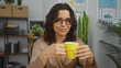 © Krakenimages.com - A beautiful brunette hispanic woman in an office setting holding a yellow cup, dressed in a beige blouse with a workplace background featuring shelves, plants, and charts.