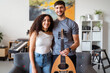 © Studio Marmellata - man holds an oud as he stands beside a smiling woman, both posing in a warmly decorated room with various musical instruments and shelves in the background, creating a relaxed and inviting atmosphere.
