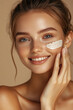 © Inspire Shots Hub - A young woman with glowing skin smiles as she applies facial cream, highlighting natural beauty and skincare on a beige background.