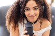 © Studio Marmellata - A woman with curly hair and a black choker smiles as she speaks into a professional microphone, seated in a cozy setting