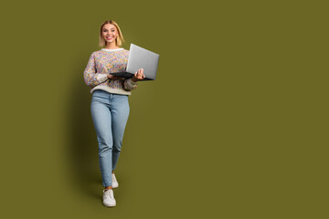 Young woman holding laptop against a khaki background, showcasing stylish knitwear and jeans