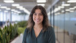 © Tom - A smiling young Hispanic woman with curly hair stands confidently in a modern office environment, surrounded by greenery and natural light, promoting a positive workplace vibe.