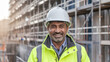 © Tom - A South Asian male construction manager wearing a hard hat and high-visibility jacket, smiling confidently on a construction site with scaffolding in the background.
