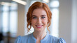 © Tom - A young Caucasian woman with red hair smiles warmly, dressed in a light blue blouse, standing in a modern, bright office setting with soft natural light streaming in.