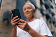 © dikushin - Selective focus of gray-haired senior lady using smartphone texting message standing on urban street on summertime. Middle aged woman grandma hold phone typing sms enjoying communication in mobile app