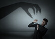 © New Africa - Young man scared by monstrous hand reaching for him. Creepy shadow on grey wall