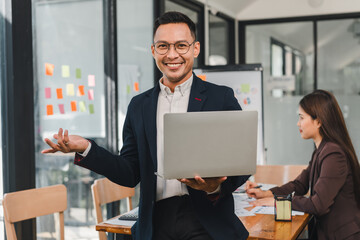 confident man in suit holds laptop while smiling in modern office. woman works in background, showcasing teamwork and collaboration