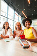 © Jose Calsina - Vertical. Three diverse happy young women friends smiling while making contactless mobile payment with smartphone and credit card terminal at modern cafe during coffee meeting time together