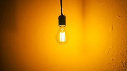 A single incandescent light bulb hangs against a vibrant yellow textured background, illuminating the space with a warm glow.