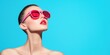 © Kamonwan - A stylish model poses with oversized pink sunglasses against a vibrant blue background, showcasing a modern and fashionable look.