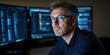 © Kamonwan - A focused individual wearing glasses sits in front of multiple monitors, immersed in programming or data analysis in a dimly lit environment.