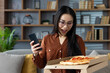 © Liubomir - Asian woman at home holding phone excitedly receiving pizza delivery. Captures moment of joy, surprise, and indulgence as pizza is revealed, highlighting fun and technology connection.