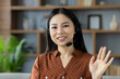 © Liubomir - Cheerful Asian woman wearing headset, participating in online communication from comfortable setting, waves with friendly gesture. Image captures remote work, technology, connection, comfort