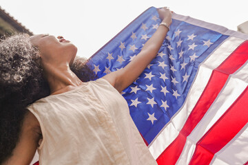 Smiling young woman holding an American flag with arms outstretched, radiating happiness and pride. The flag's bold colors contrast with her serene expression, symbolizing freedom and positivity.