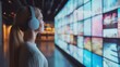 © thenort - Young woman wearing headphones looks at a wall of screens displaying images and videos.