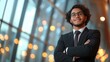 © familymedia - A young professional in a suit confidently stands with folded arms in a modern office space, illuminated by soft, ambient lighting through large windows.