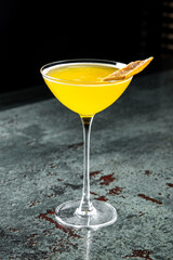 Wall Mural - A vibrant yellow cocktail in a classic martini glass, garnished with a twist of citrus peel, set against a dark background with a textured surface. Perfect for a and elegant drink presentation.