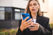 © zvkate - Serious pensive business leader woman getting message on smartphone and thinking. Young woman holding mobile phone and look doubtful. Girl wear jacket and stand near office in the city.