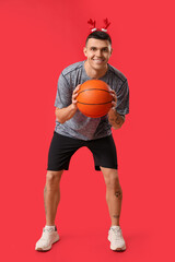 Sticker - Young happy man in reindeer horns playing basketball on red background