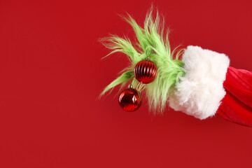 Green hairy hand of creature in Santa costume with Christmas balls on red background