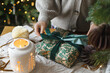 © sonyachny - Merry Christmas and Happy holidays! Hands in cozy sweater wrapping stylish christmas gift on table with glowing lantern and modern paper ornaments against golden tree lights. Atmospheric eve