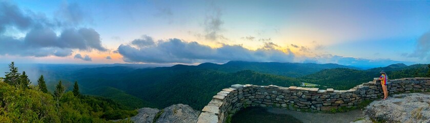 Wall Mural - Woman standing at a viewpoint gazing at majestic mountains during sunset