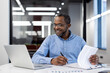 © Liubomir - Mature African American businessman analyzing reports in modern office. Professional man working on laptop, reviewing financial documents with focus, confidence, and a smile on his face.
