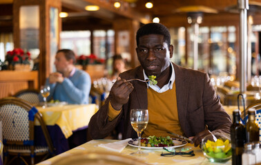 Adult african american man enjoying delicious lunch in cozy restaurant, sitting at served table and eating fresh vegetable salad