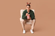 © Pixel-Shot - Young man with bucket of popcorn watching movie and sitting in armchair on brown background. Christmas celebration