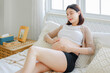 © Auttapol - Young Asian happy pregnant woman is sitting and relaxing on bed and and touching her belly. Family plan, pregnancy, motherhood, people and expectation concept