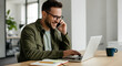 © Paintbrush - A man in glasses smiles while talking on the phone and working on a laptop in an office.