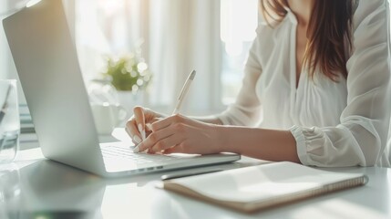 Fototapete - A woman writing notes while working on a laptop in a bright, airy workspace, surrounded by natural light.